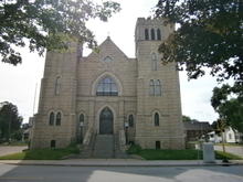 Two-story, Gothic building in light colored brick with central entry flanked by four-story corner towers. 